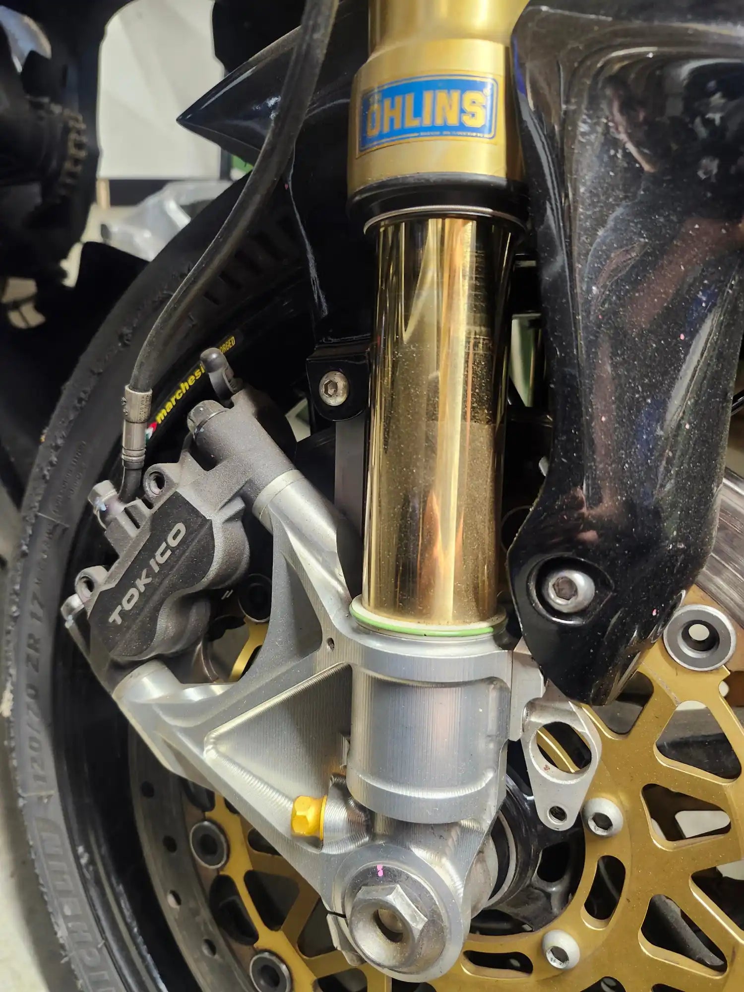 Fourche avant de moto Öhlins en or avec l'étrier de frein et le disque de frein visibles.